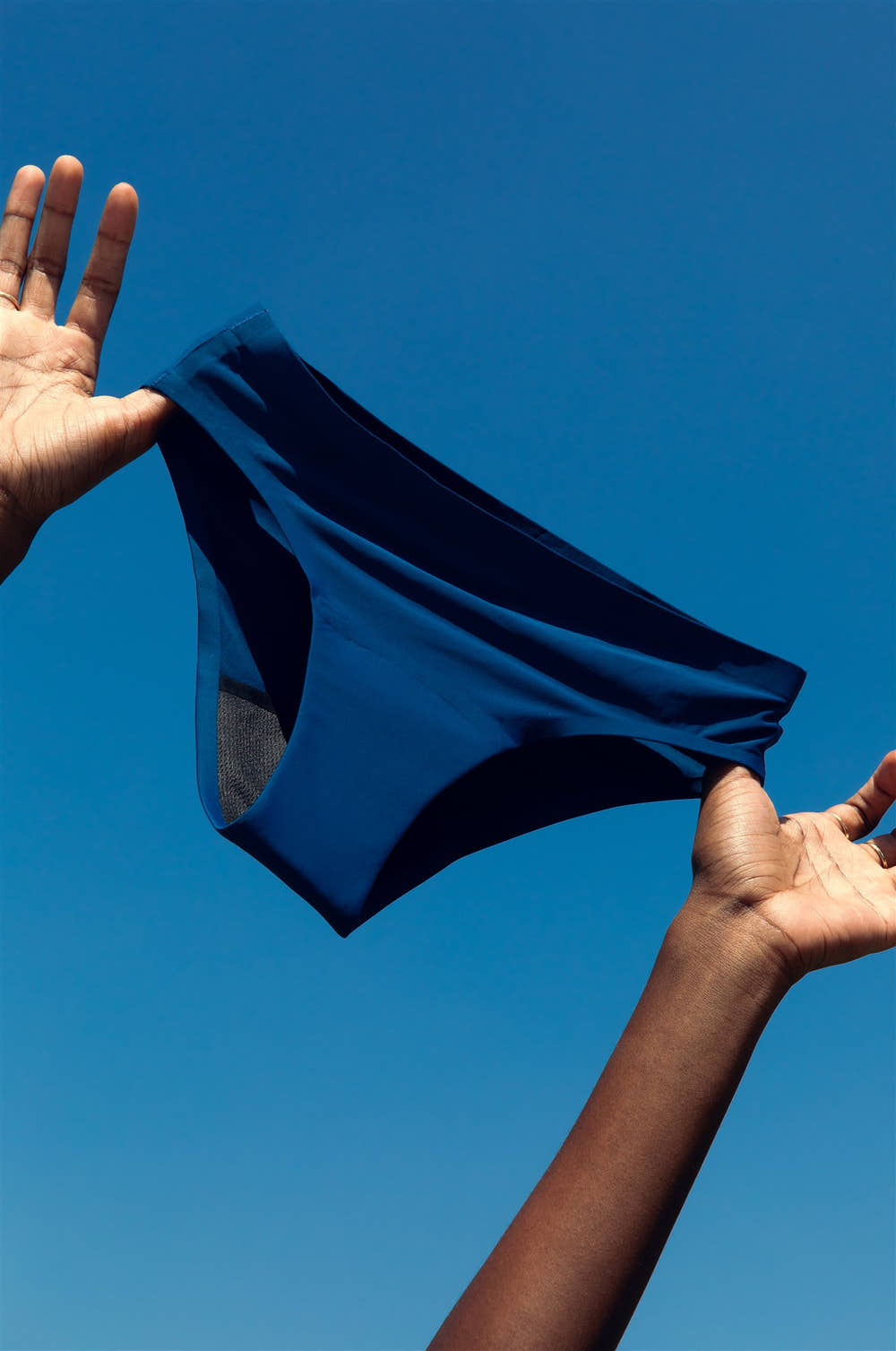 Hands holding up SweeTee navy blue period underwear against a blue sky, showing built-in absorbent liner